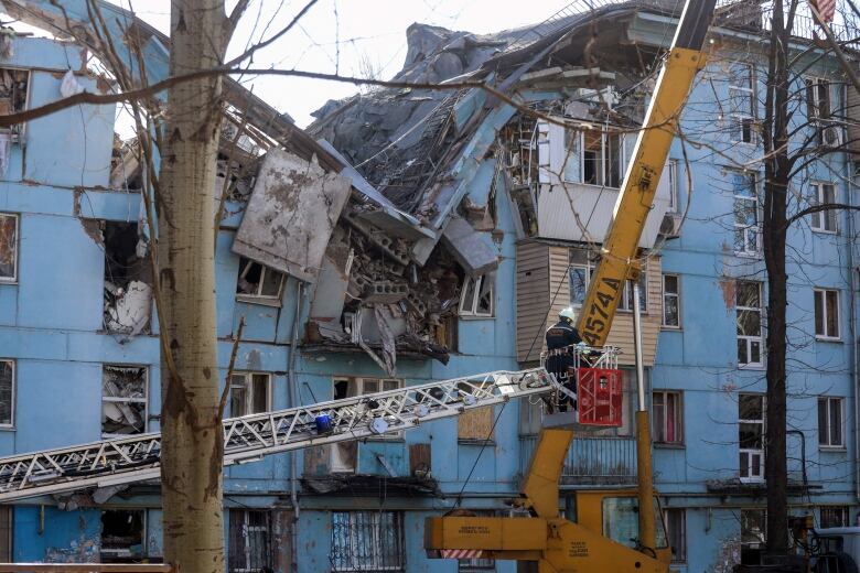 A first responder in a bucket lifted by a crane is shown in front of a heavily damaged residential building.