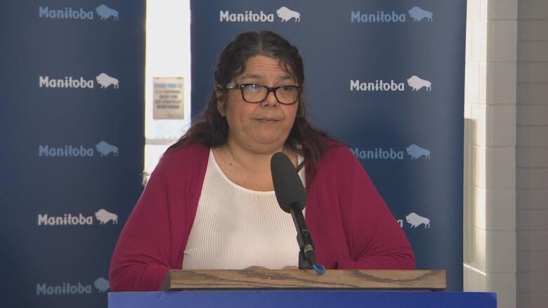 A woman wearing glasses, red cardigan and white shirt speaks into a microphone at a news announcement.