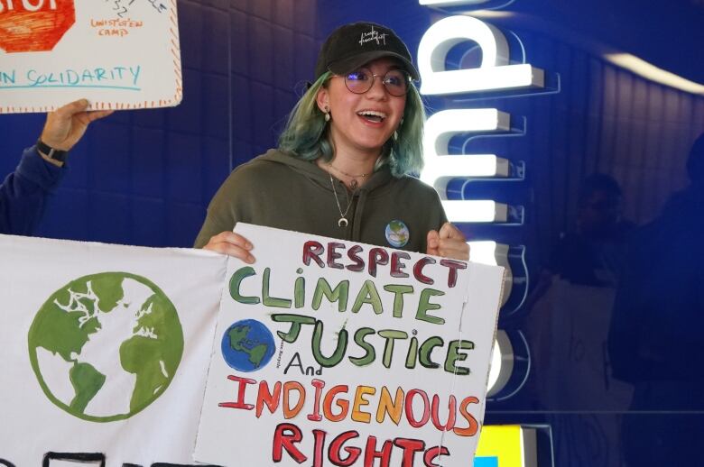 A young woman with a baseball cap and glasses holds a sign that reads, 