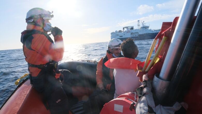 Two men wearing white helmets and orange jackets lean on the edge of a boat, as they talk with another man, while adrift on the sea. Behind them, a larger boat is seen.