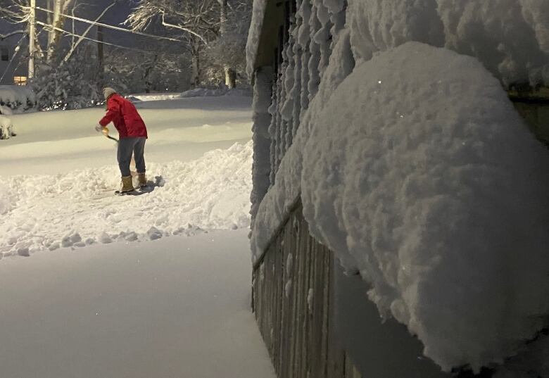 A person in a red jacket is seen shovelling snow off a driveway in the early morning hours.