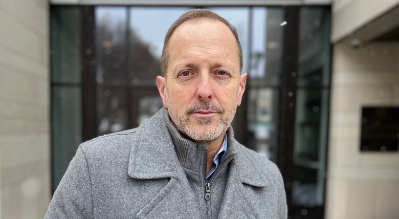 A man in a grey coat and with a serious expression on his face stands outside a building looking directly at the camera.