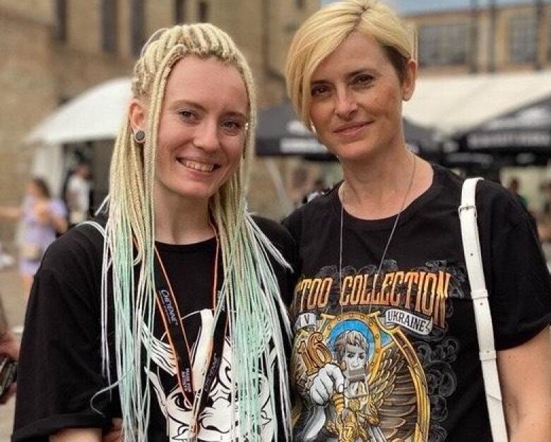 Two women, dressed in black T-shirts, one with long blonde hair and another with short blonde hair, smile and look straight on to the camera. 