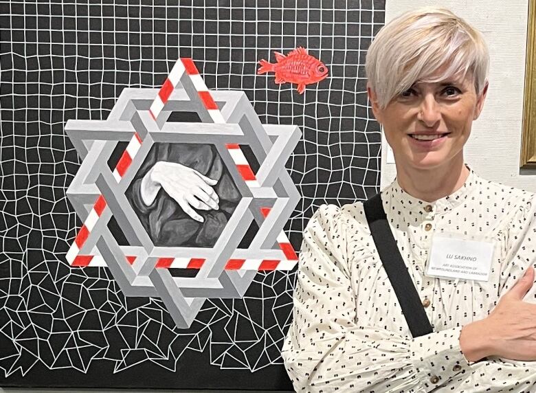 A woman poses with her arms crossed next to a piece of art that is black and white squares with a multi-shape figure in the centre with a white and red and white triangle. 