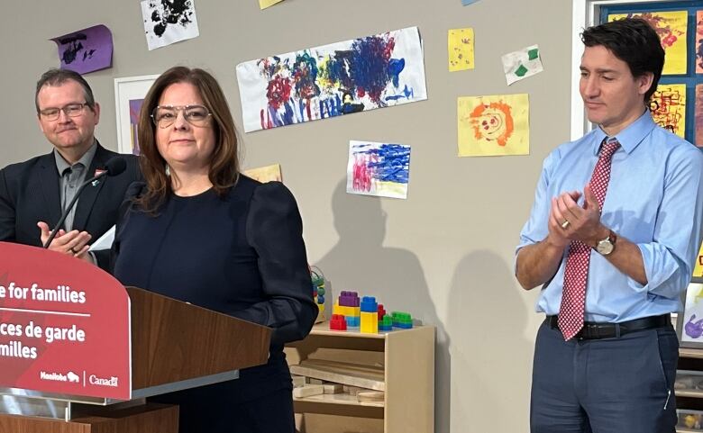 A woman with long hair and glasses smiles as she stands at a podium. A man in short black hair applauds behind her.