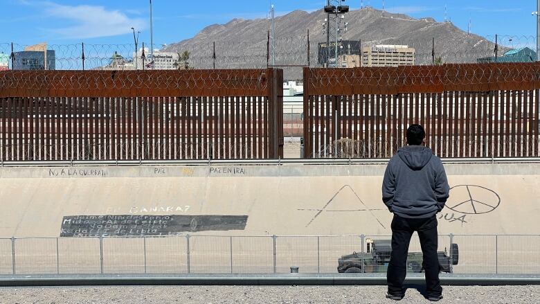 A man wearing black jeans and a grey hooded sweatshirt stands in front a cement barrier place in front of a metal and barbwire fence. 