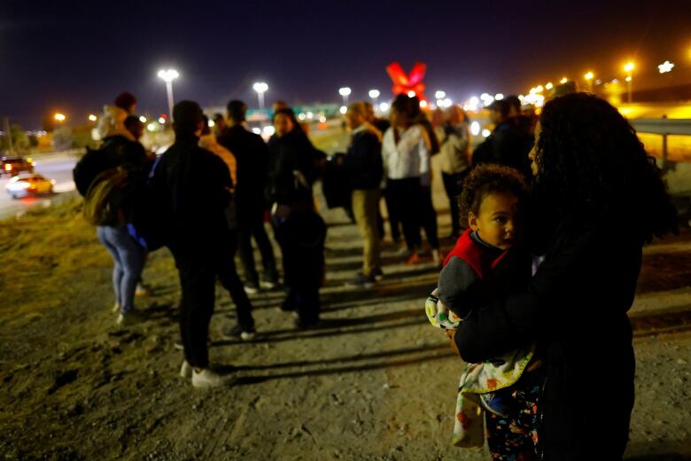 A group of people stand scattered in the dark in the dirt next to a roadway, with streetlights illuminated in the background.