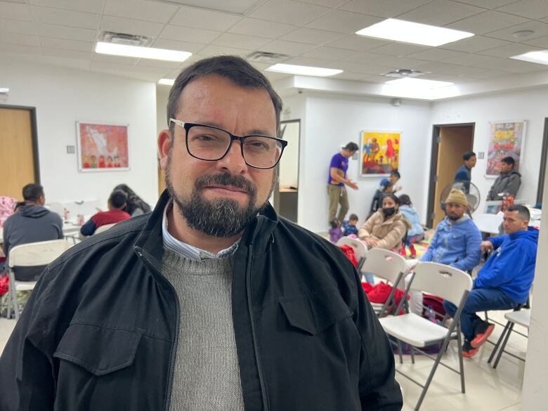 A man with black rim glasses and a beard, wearing a grey sweater and black jacket, poses for a photo in the waiting room of a building.