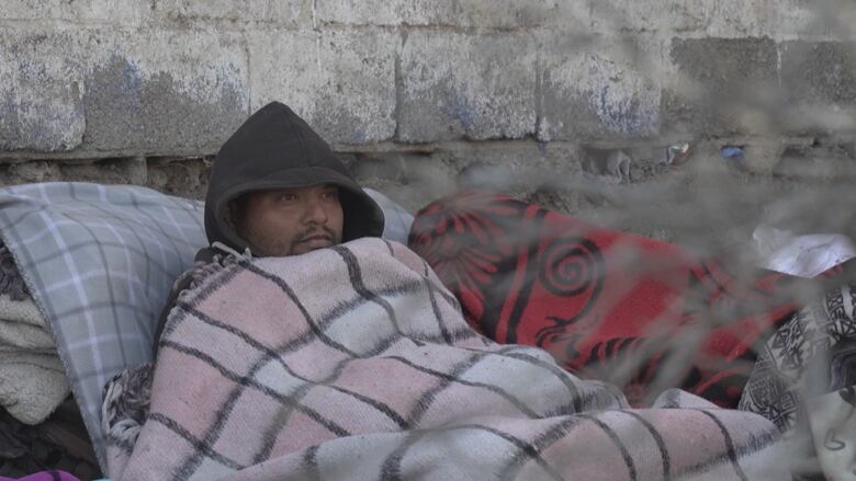 A man wearing a hooded jacket huddles until a white, pink and black plaid blanket, leaning up against a cinder-block wall. 