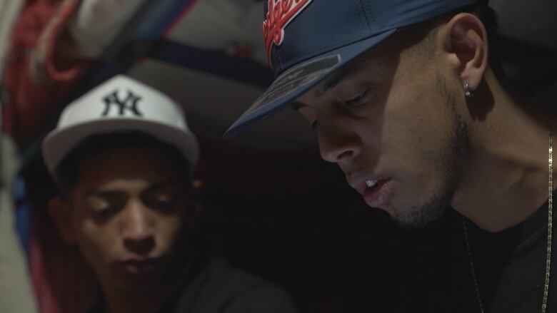 The faces of two young men in baseball hats are illuminated by the light from a cellphone screen, as they sit on the bottom bunk of a bunkbed.