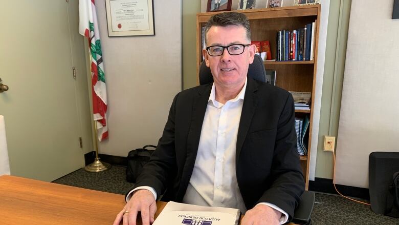 A man in a suit jacket sits at a desk in an office with a report on the desk in front of him.