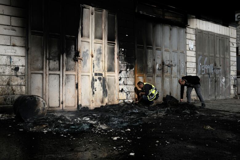 Two men, one wearing a reflective yellow vest, stoop near rubble outside a smoke-blackened building to examine damage. 