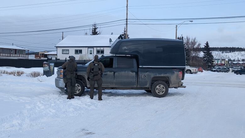 Two police officers in tactical gear stand behind a vehicle on a snowy residential street.