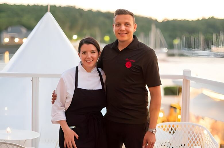 A woman in a chefs apron stands next to a man in black.