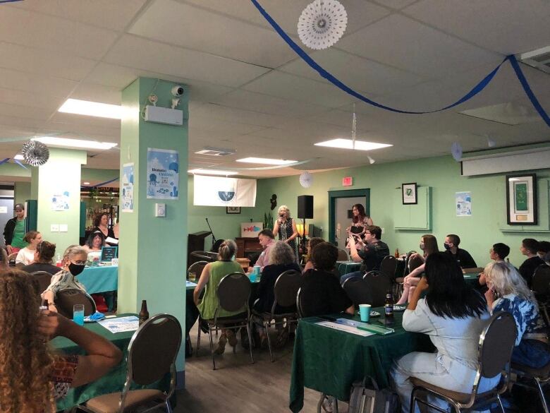 A large group of people are gathered in a green room filled with tables and chairs. A woman is speaking at a microphone.