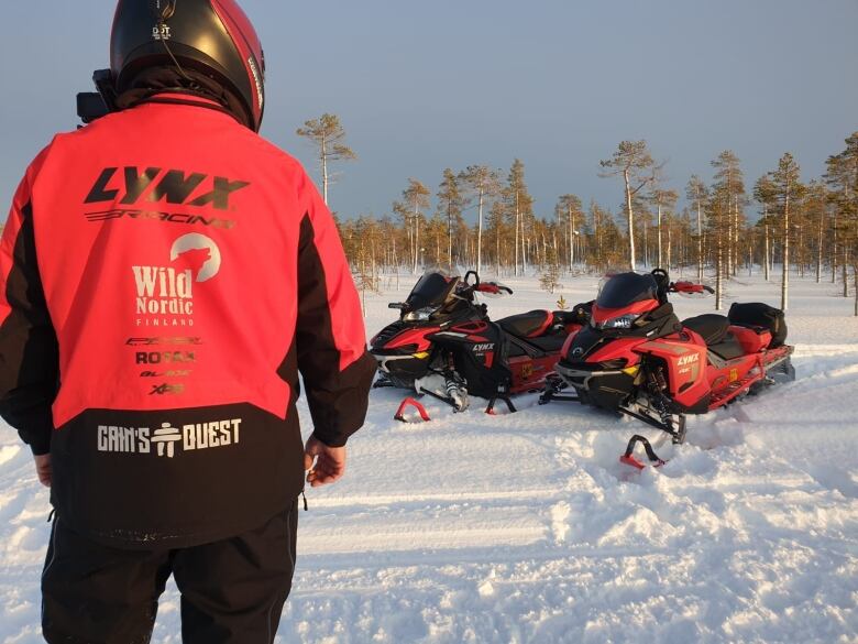 A in a red snowmobile jacket turned back on to the camera with two Bombardier Lynx snowmobiles in the background.