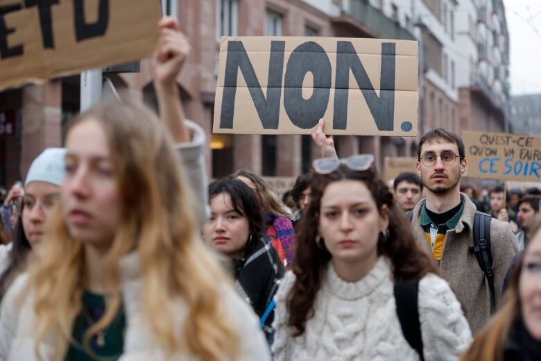 A number of young people, one with a sign saying 'Non,' are shown walking in a street.