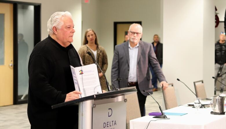 A man wearing a black shirt and pants standing at a podium holding a folder with documents as others listen in the background of a conference room. 