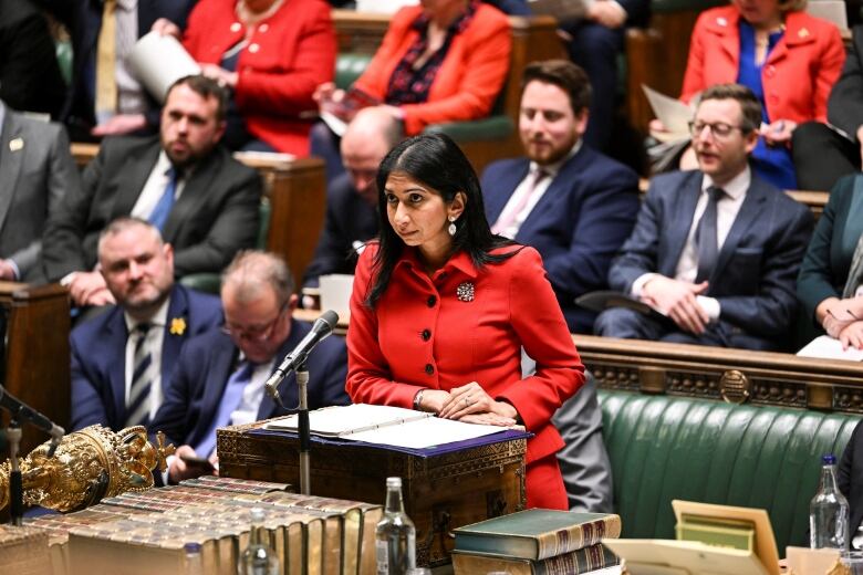 A woman is seen speaking at a podium inside a legislative chamber that appears well populated.