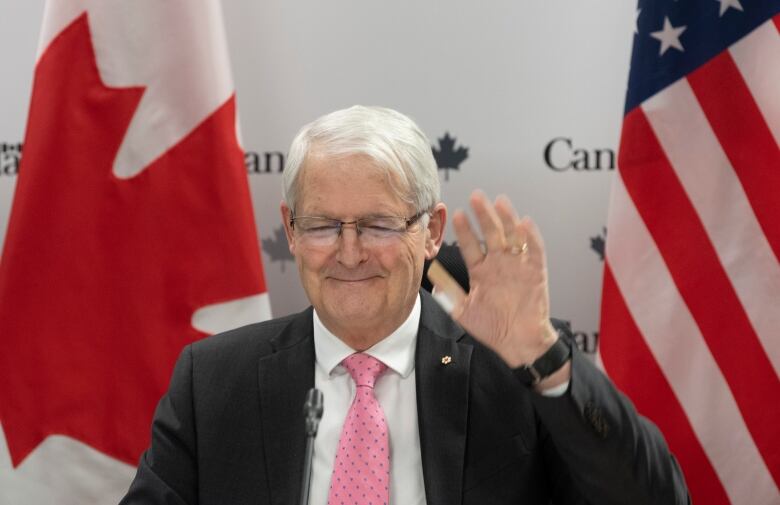 A man with gray hair wearing a suit with a pink tie waves and smiles.