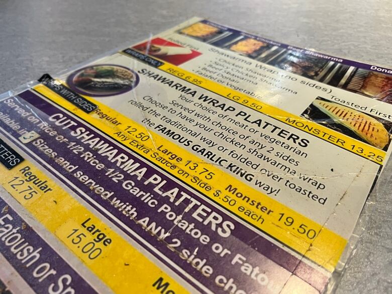 A beat-up looking menu in purple and yellow shows food prices. Some prices are covered in stickers to change them.