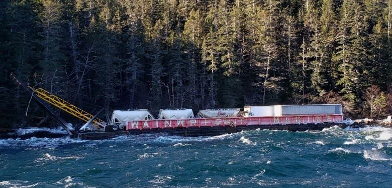 A red barge loaded with white containers is pictured after running aground in front of an evergreen forest.