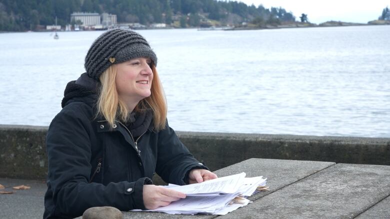 A woman sits at a picnic bench by the seashore holding paperwork.