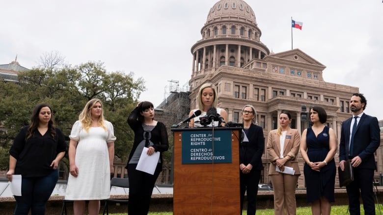 A woman speaks into a mic at a podium outside, as seven other people stand behind her.