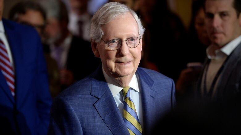 A man with grey hair and glasses, wearing a blue suits stands in a crowd smiling.