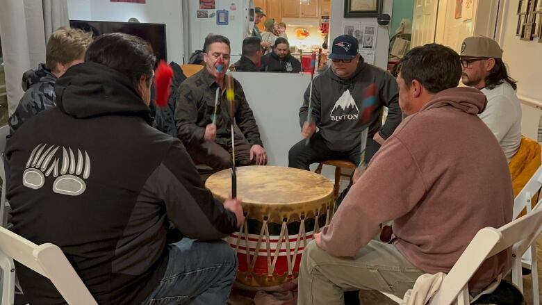 A group of men sit around a drum, each one holding a drum stick. There's an empty chair with a drum stick on it.