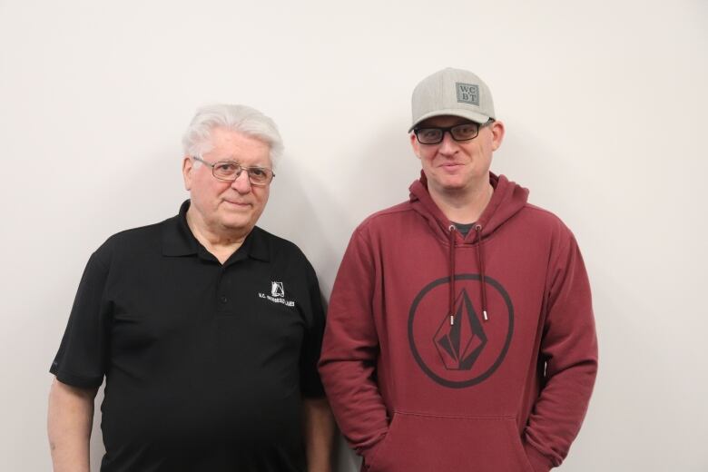 A man in a black bowling shirt stands next to a man with a beige cap and red hoodie