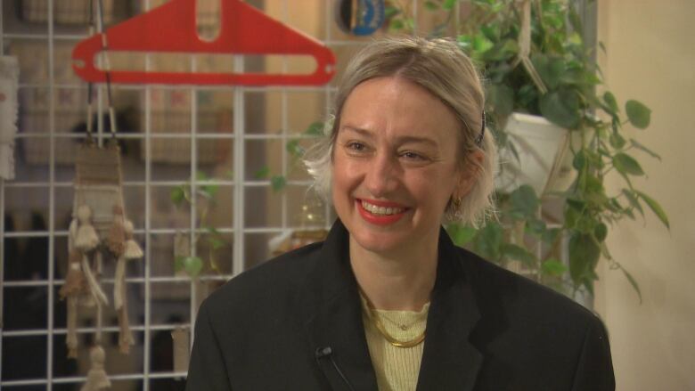 Woman with ear length hair smiles at the camera. She is wearing black blazer, yellow shirt, gold jewelry and red lipstick.