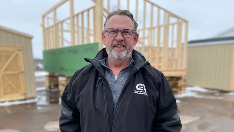 A man in a black jacket stands in front of a tiny home under construction 