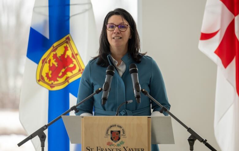 A woman with long dark hair and glasses stands behind a podium with flags in the background.