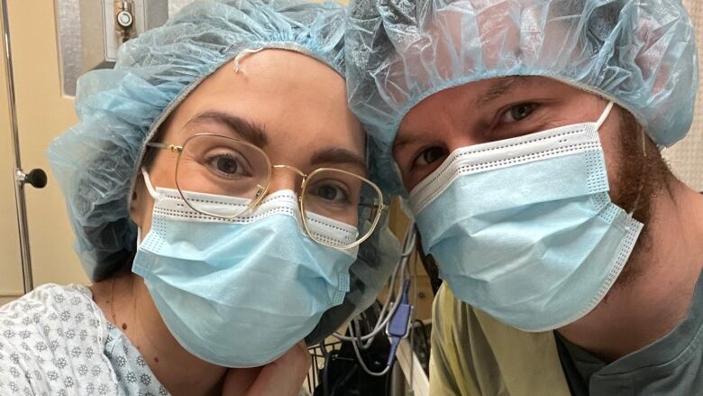 A man and woman are shown in a health care setting wearing blue face coverings and plastic covers over their hair.