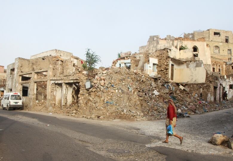 A man is shown walking in sandals on a street, with a damaged building and rubble shown on the street corner.