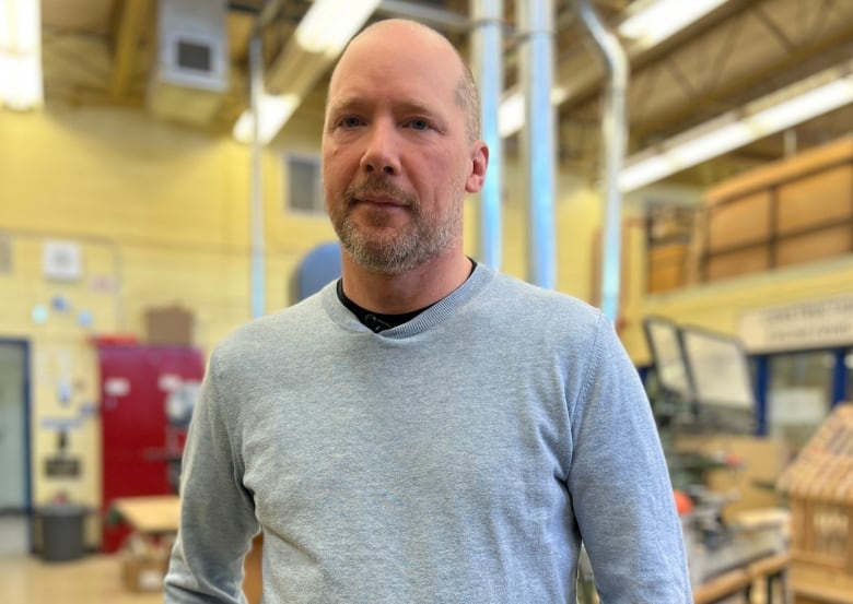 A man in a light crewneck sweater stands in a large high school shop classroom. 