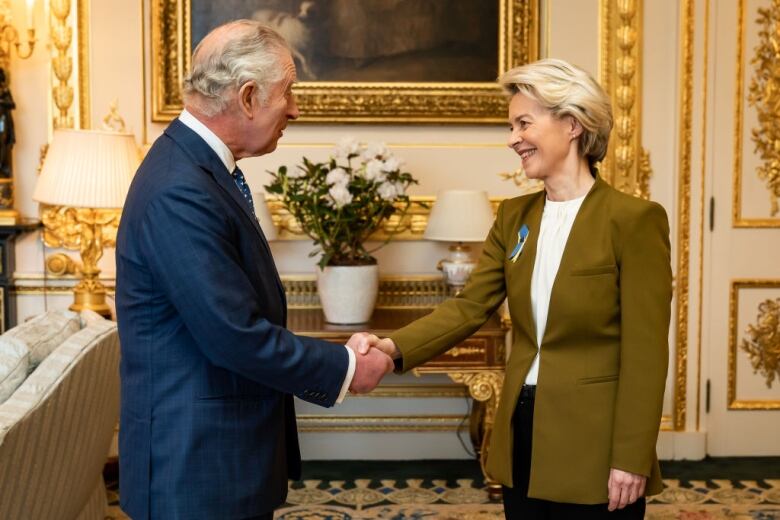 Two people shake hands in front of a table with flowers on it and a portrait hanging on a wall.