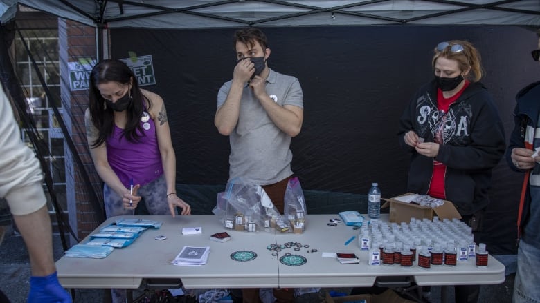 Three people wearing facemasks stand at a table, with medical paraphernalia and hand sanitizers visible on it.