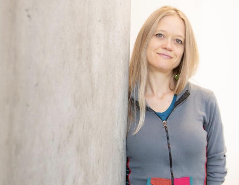 Dames smiles at camera leaning on a concrete wall with long blonde hair and wearing a grey sweatshirt. 