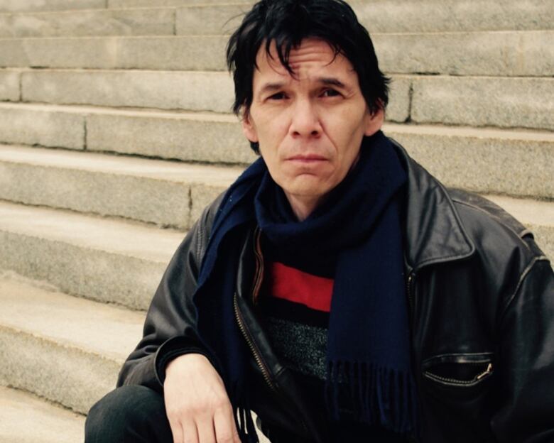 Man with jet black hair sits outside in black leather jacket, jeans and navy scarf with red detailing. He stares at the camera intently. 