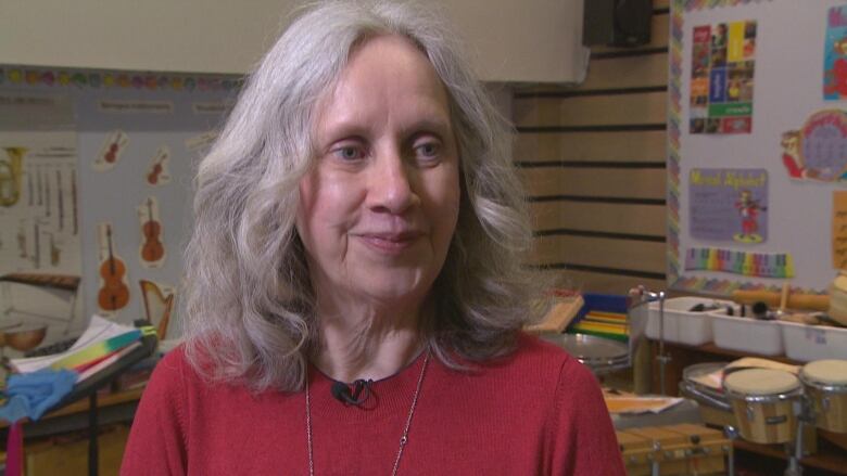 Woman with grey hair smiles at camera. She is wearing a red sweater and is in a music classroom.