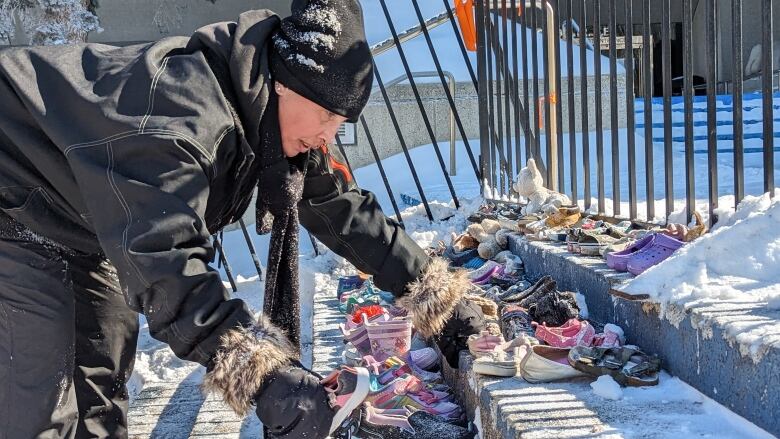 a person dressed in a black jacket, toque and mittens leans down on concrete steps to rearrange a lineup of children's shoes