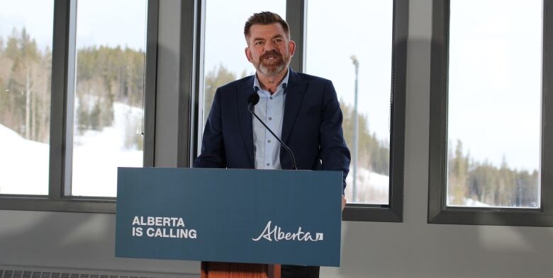 A man wearing a blue suit and light blue shirt stands and speaks at a podium that has a sign that says Alberta is Calling.