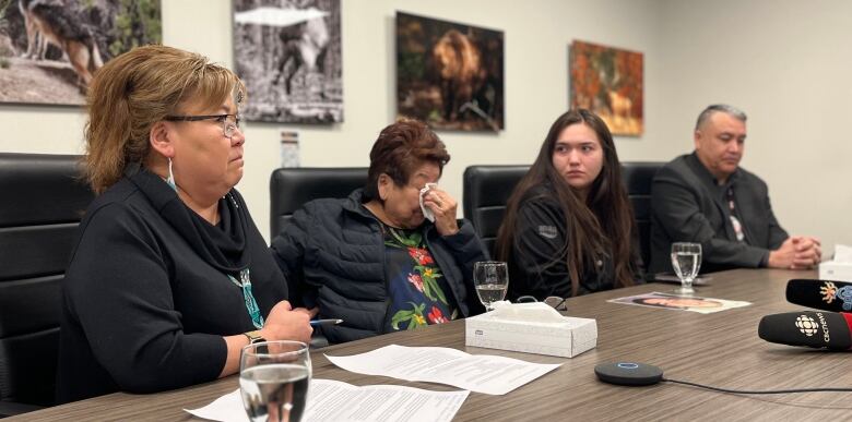 Four people are seated at a conference room table at a media briefing. An older woman dabs her eye with a tissue at centre.