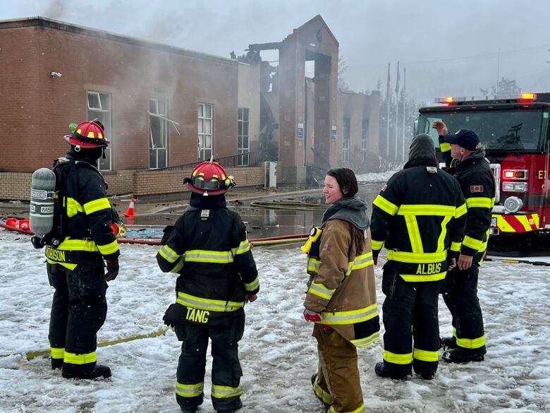 Several firefighters stand in front of a burned building with smoke coming out of it. 