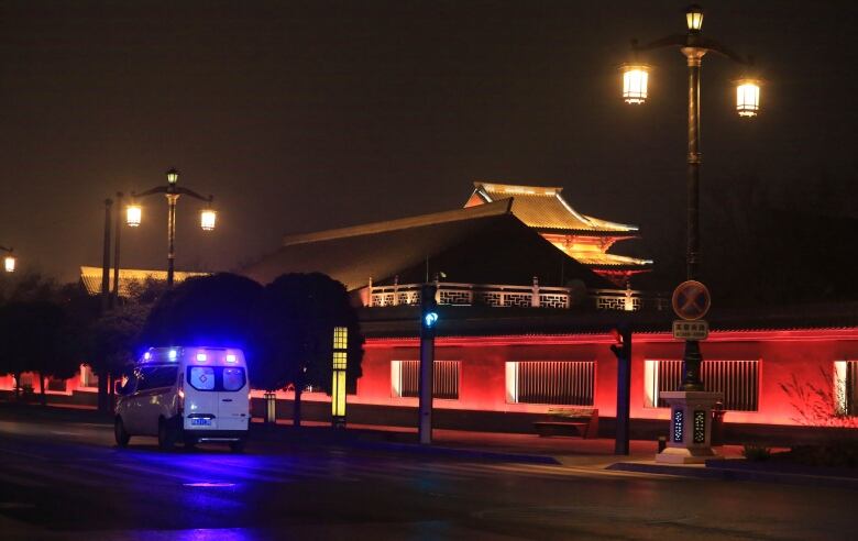 An ambulance with its lights on faces away from the camera on a wet, paved street. Street lights, a large red barrier wall and Chinese architecture can be seen in the distance.