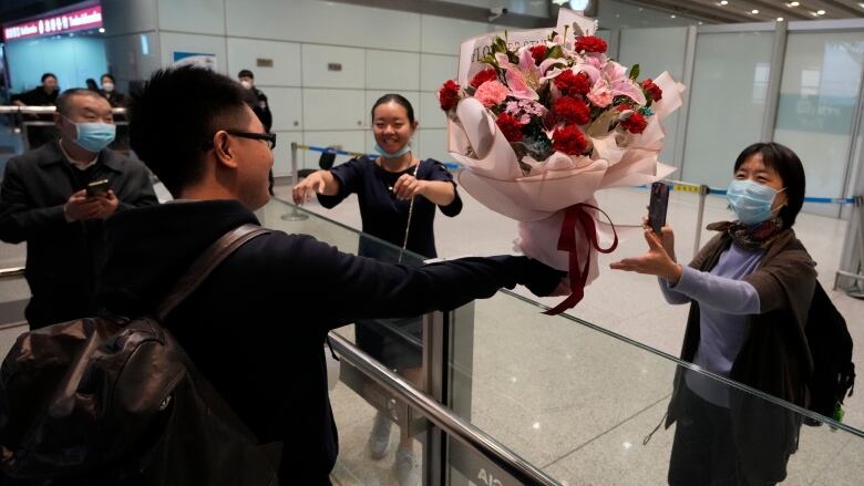 A man wearing glasses reaches a large bouquet of red and pink flowers over a glass barrier to two women who are both outstretching their arms.