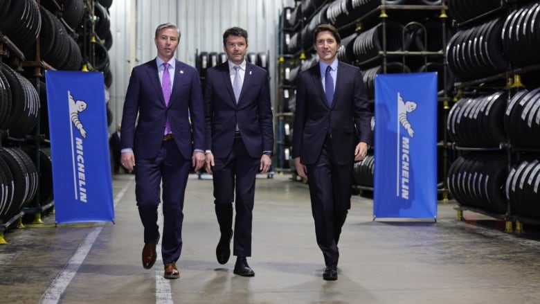 Three men in suits walk in a factory flanked by tires.