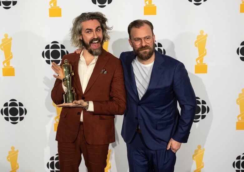 Two men smile and pose for a photo. The man on the left holds up a statuette.